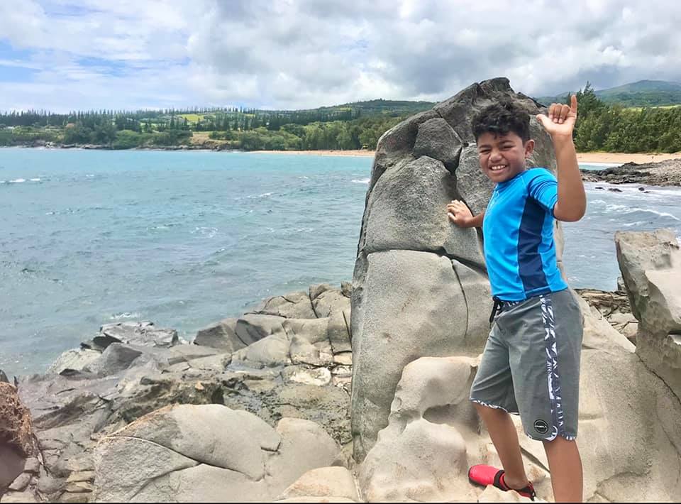 A young boy stands near a large rock with a hang ten sign in Maui.