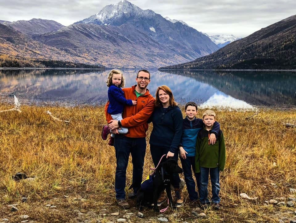 A family of five with their dog stands on a grassy area with a glacial lake and mountains behind them.