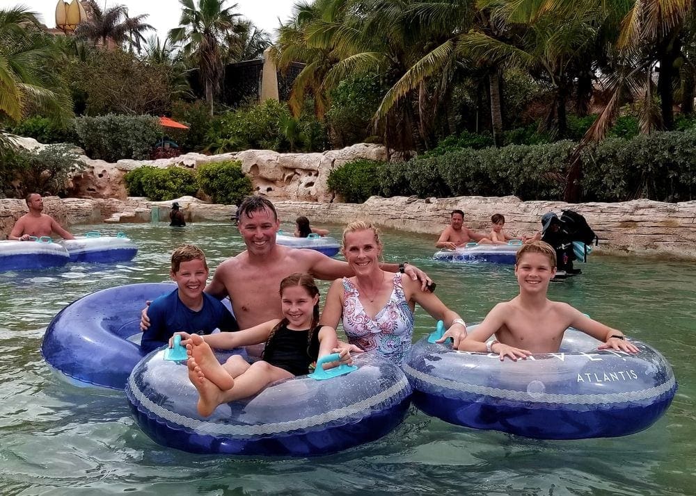 Three kids sit in tubes, while their parents stand behind them in a lazy river.