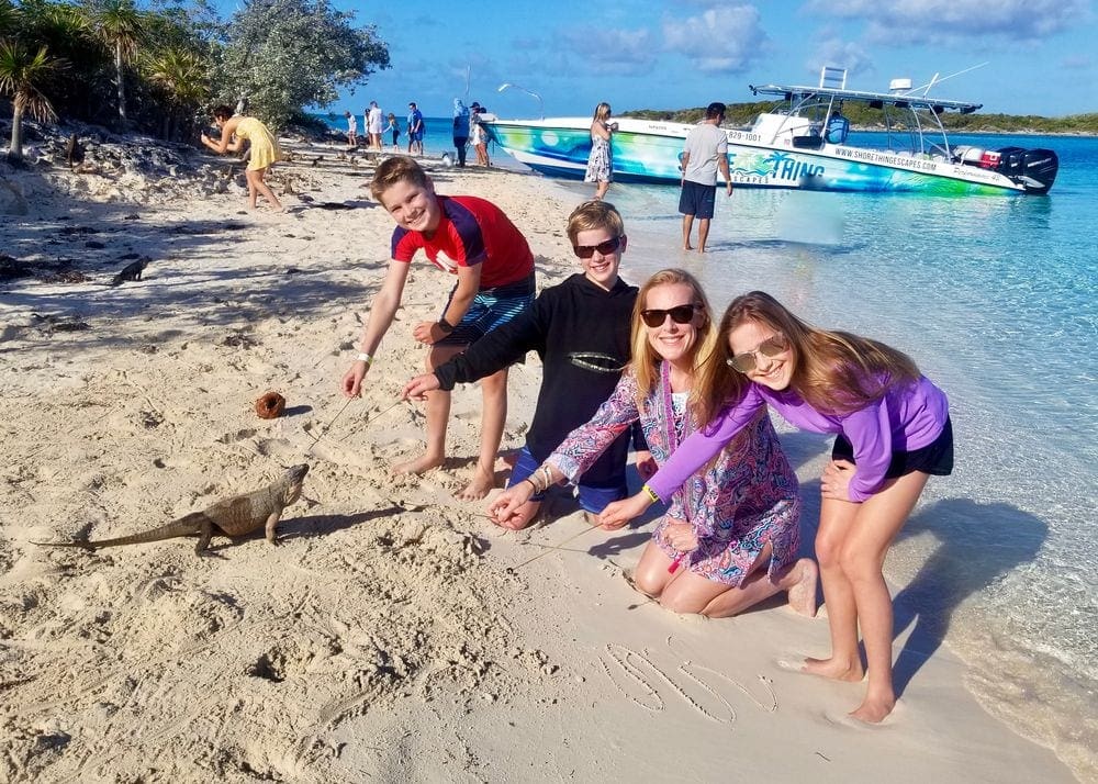 Mom and three kids feeding an iguana with a wooden skewer on a beach in the Bahamas.