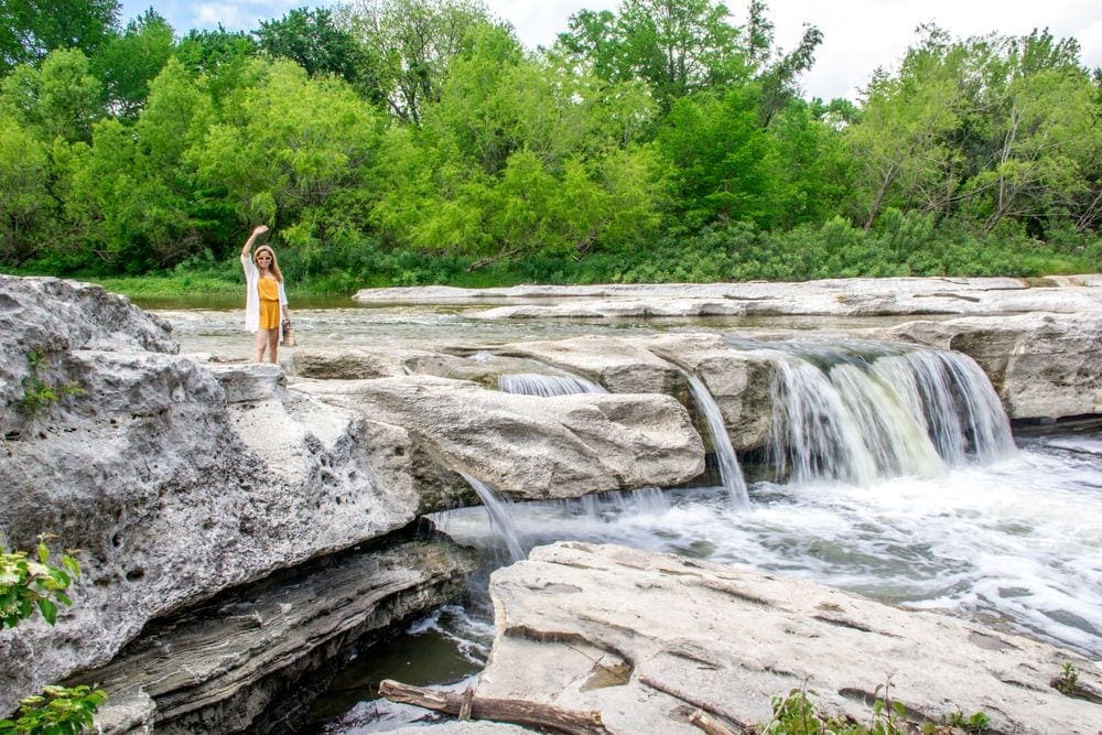 A woman in yellow waves from atop the falls at the McKinney Falls State Park.