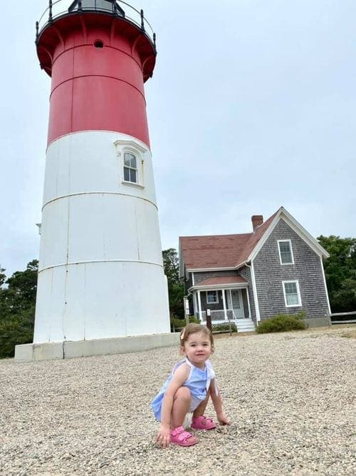A toddler girl crouches in front of a lighthouse in Cape Cod.