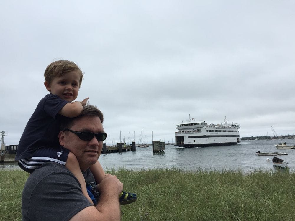 A young boy rides his dad's houlders with the Martha's Vineyard ferry in the water in the background.