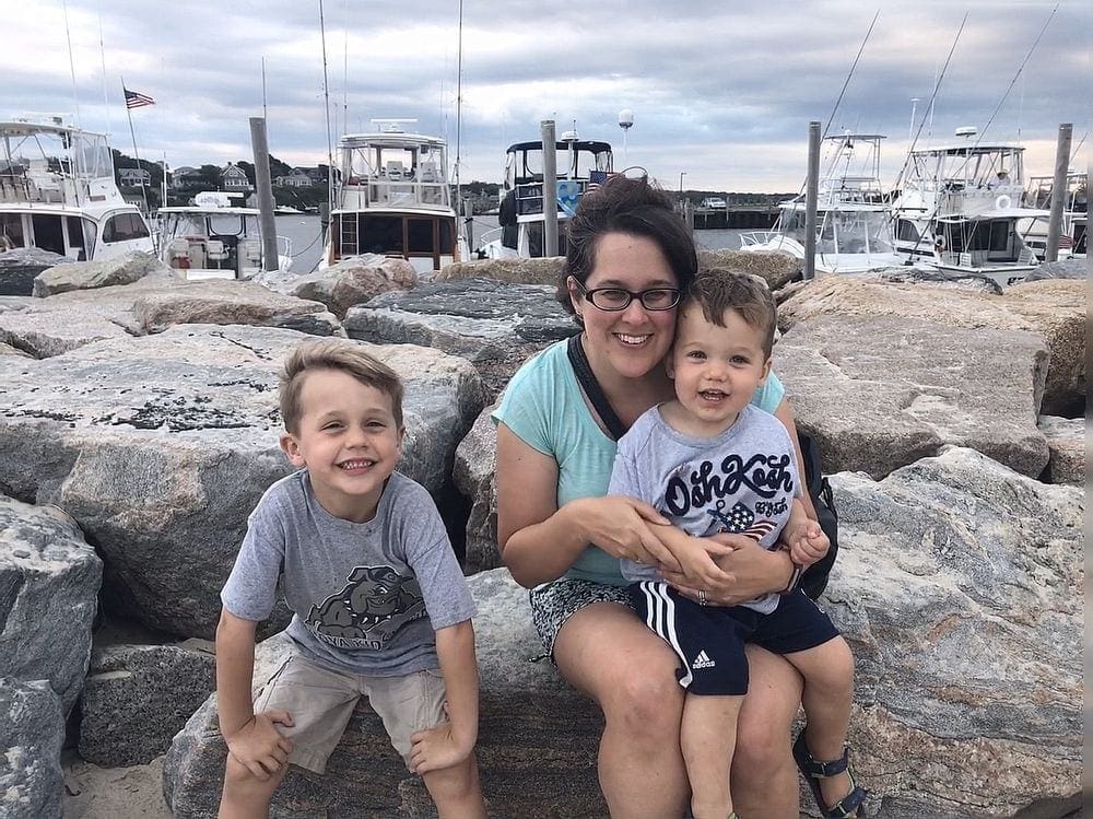 A mom sits on large rocks with her two boys with a variety of ships in the marina behind them.
