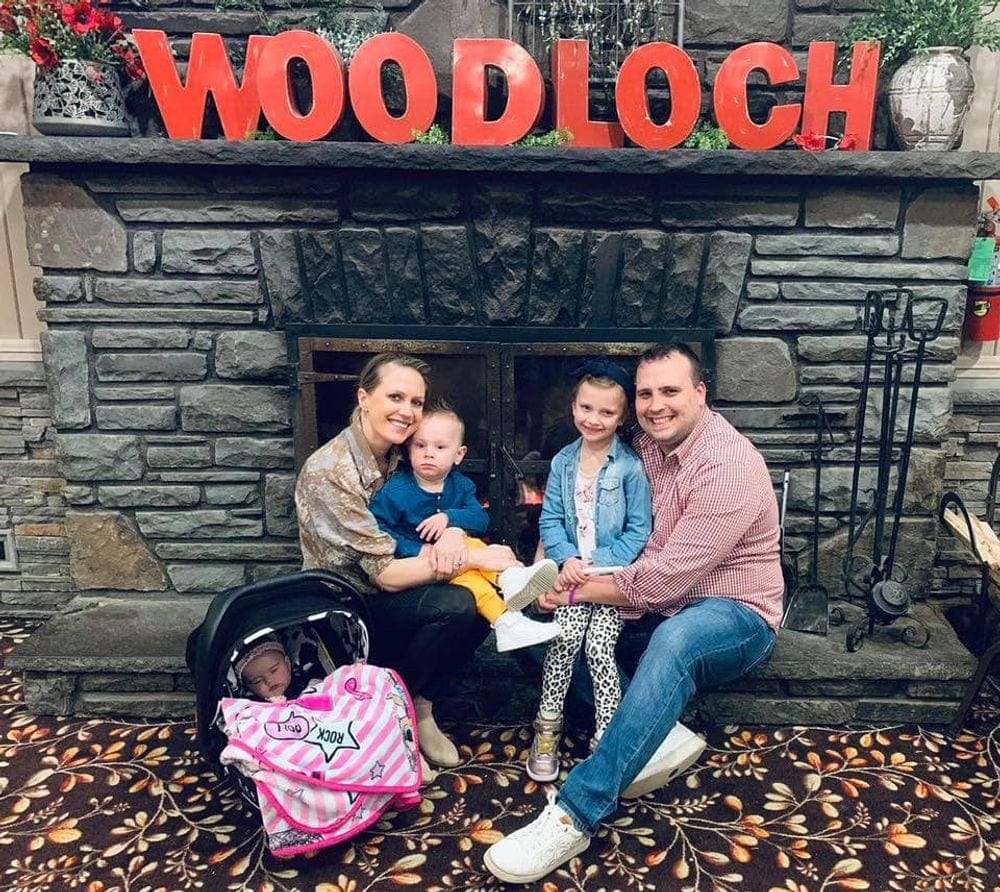 A family of five sits on the side of a fire place at Woodloch, with a large red sign overhead reading "Woodloch".