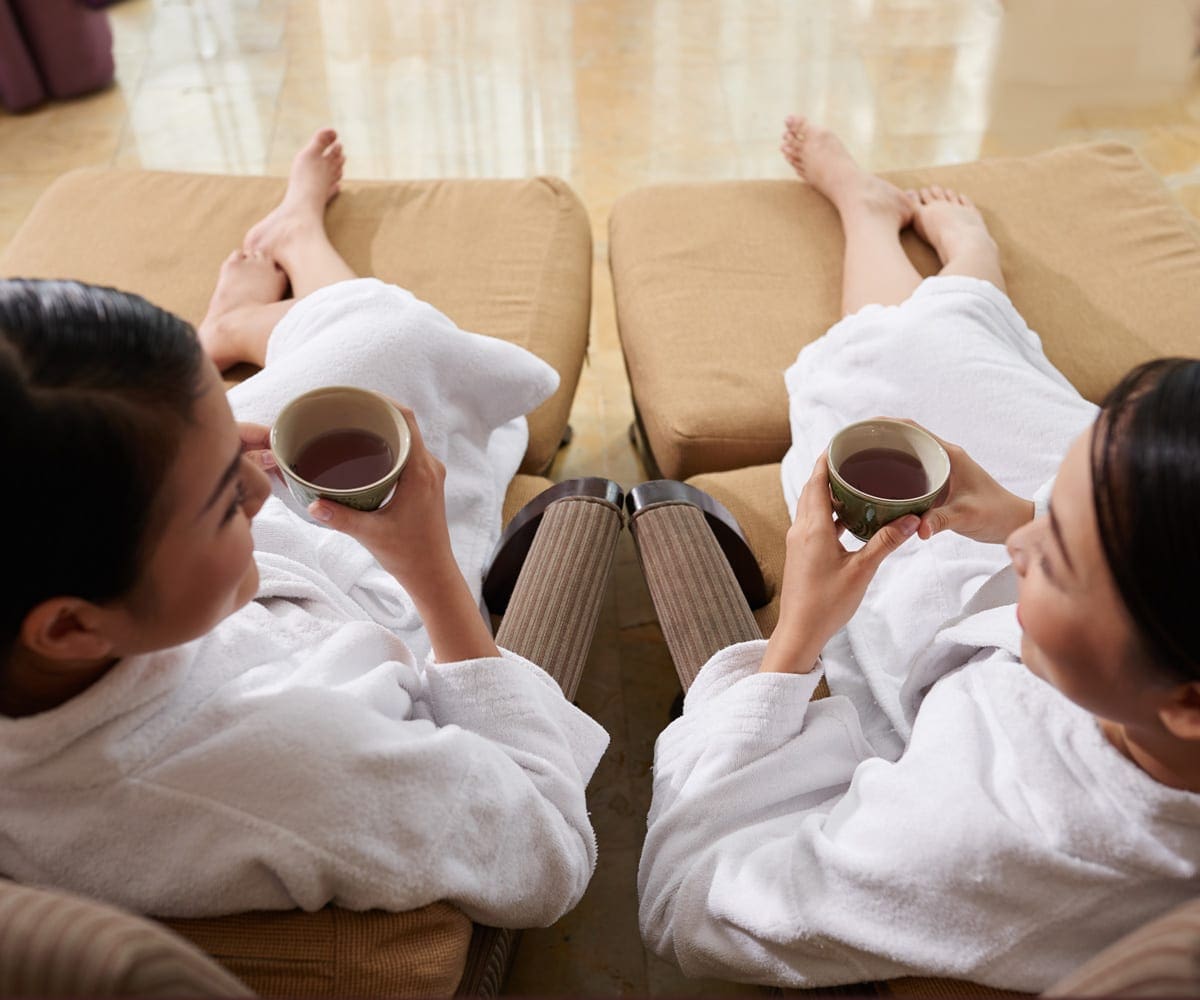 Two moms enjoy a cup of coffee while sitting in robes on spa loungers.
