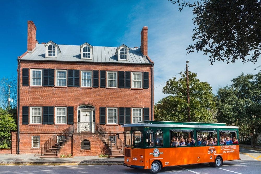 A trolly for the Old Town Trolley Tours filled with people stops outside of a large historic home in Savannah.