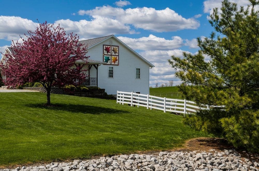 A large white building, featuring a colorful quilt sqaure, stands proudly surrounded by blooming trees on a sunny day in Shipshewana, a great stop for a Midwest road trip with kids.