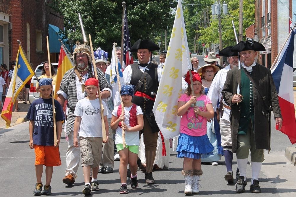 A series of parade walkers wander down a street, featuring men in colonial costumes and children carrying flags in Ste. Genevieve, a great stop for a Midwest road trip with kids.
