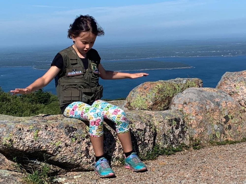 A young girl sits along a rock ledge holding our her arms as she feels the wind atop Cadillac Mountain in Acadia National Park.