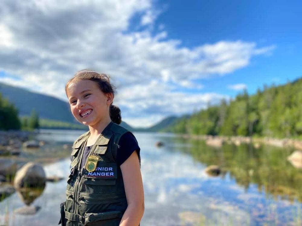 A young girl smiles broadly on a sunny day while explore Jordan's Pond in Acadia National Park, one of the best cool-weather destinations in the US for families.