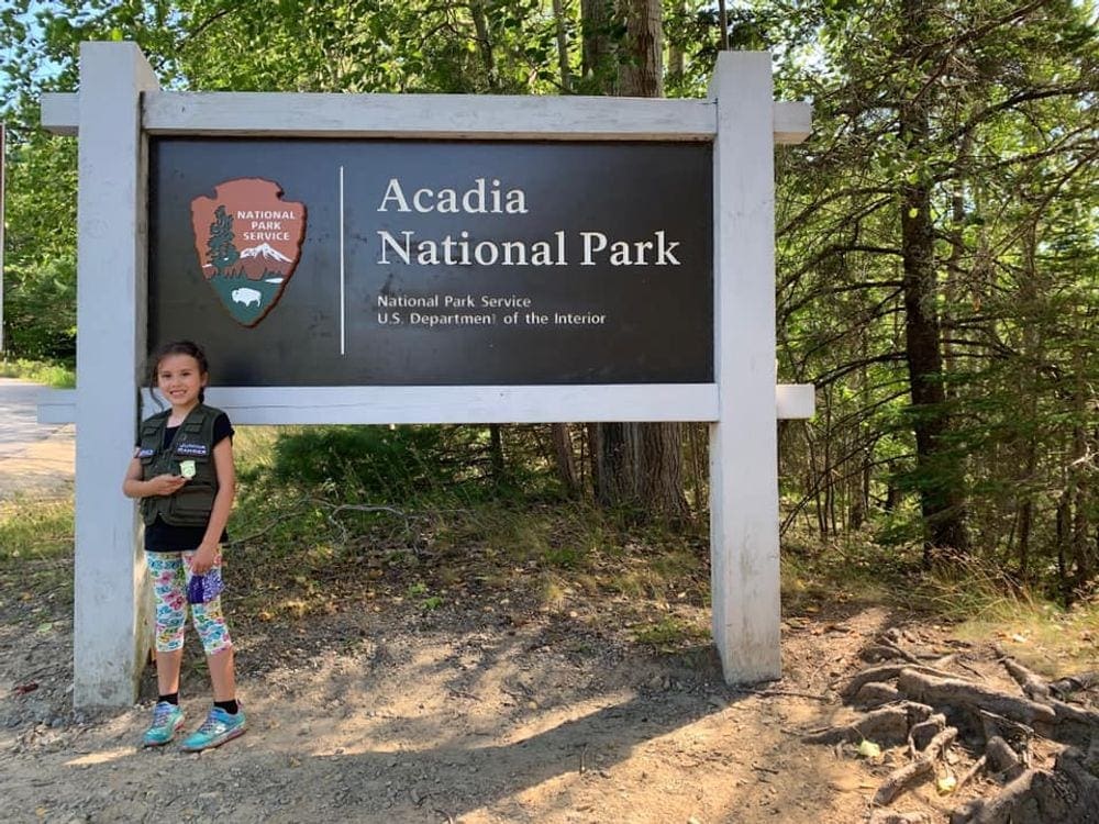 A young girl stands proudly next to the entrance sign for Acadia National Park, one of the best things to do in Bar Harbor with kids.
