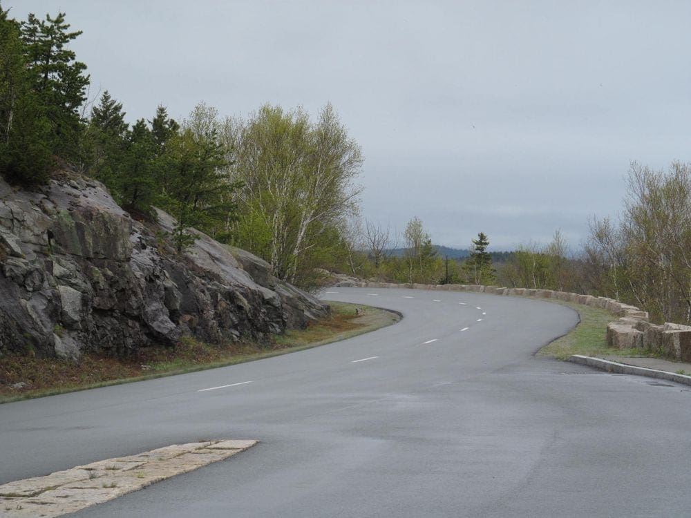 An empty road cuves to the left, with iconic Maine rocks and trees on both sides.
