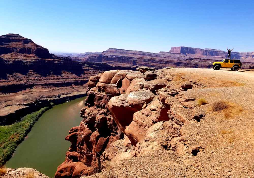 A man stands with hands in the air atop a yellow jeep with a view of a large river and cliffs inside Canyonlands National Park, one of the best things to do in Moab with kids.