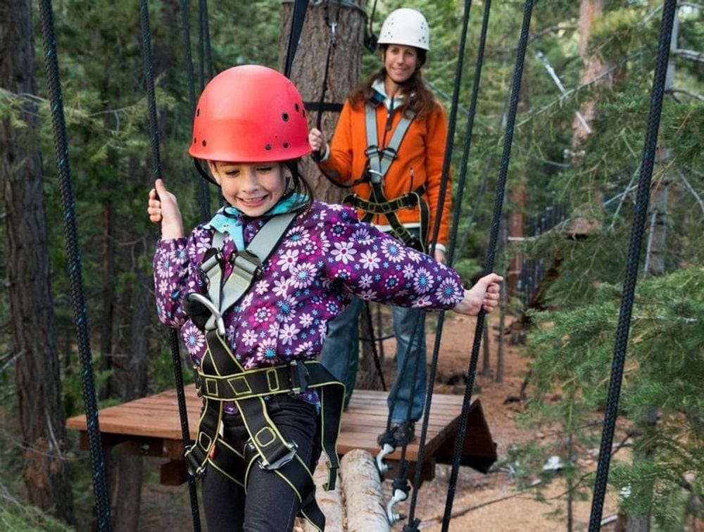A young girl makes her way across a ropes course while an adult watches on behind her.