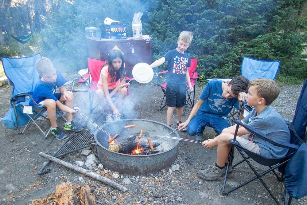 Five kids sitting in camp chairs cook hot dogs over an open fire while camping near Grand Tetons National Park.