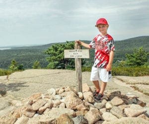 A young boy stands next to a summit sign on Bass Head Lighthouse Trail peek near Bar Harbor.