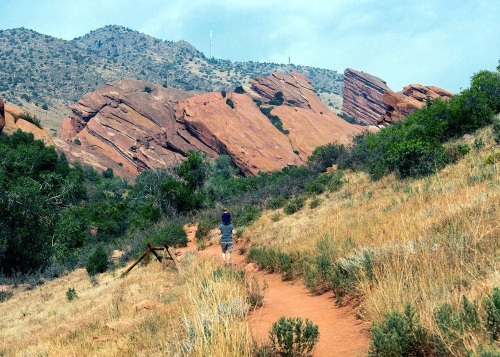 In the distance, a dad carries his young child on his shoulders while hiking at Red Rocks Amphitheater, one of the best stops on One-Week Colorado itinerary for families