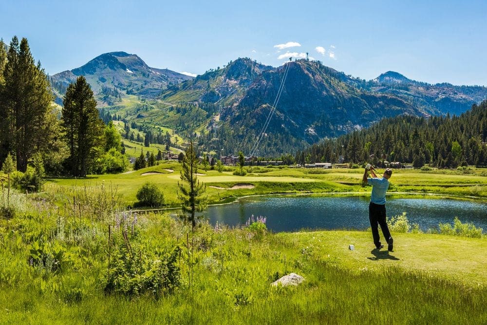 A man swings a golf club while golfing near Lake Tahoe, with mountains and a small pond in the background.