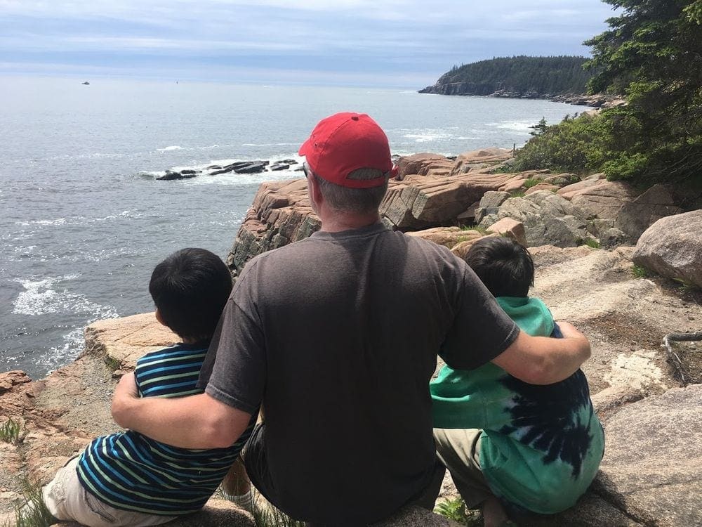 A dad holds his two young kids close while sitting atop a rock ledge in Acadia National Park, looking onto the stunning ocean view.