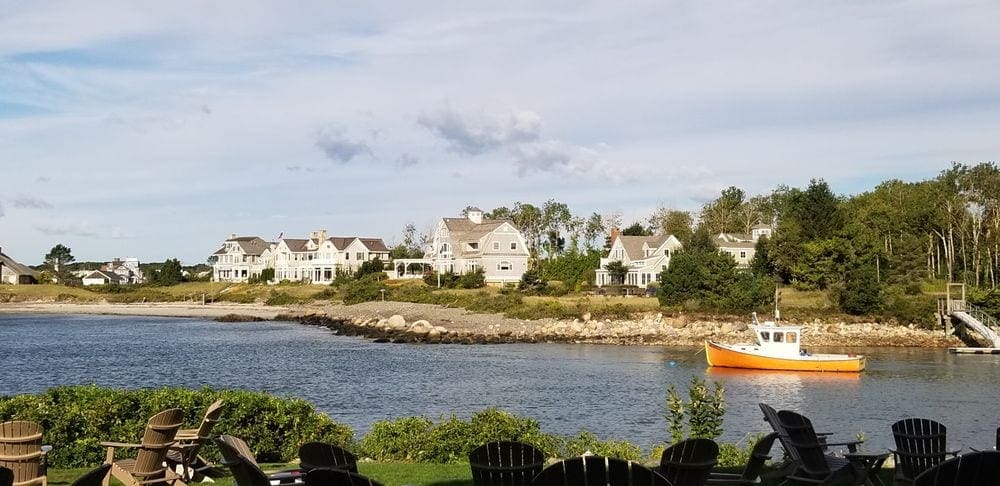 A yellow tug boat move along a bay near Kennebunkport, Maine, with classic Maine home in the background.