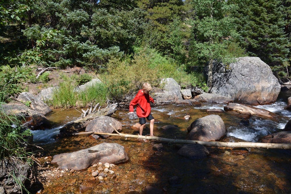 A young boy wearing a red sweatshirt balances while walking across a fallen log over a stream in Colorado.