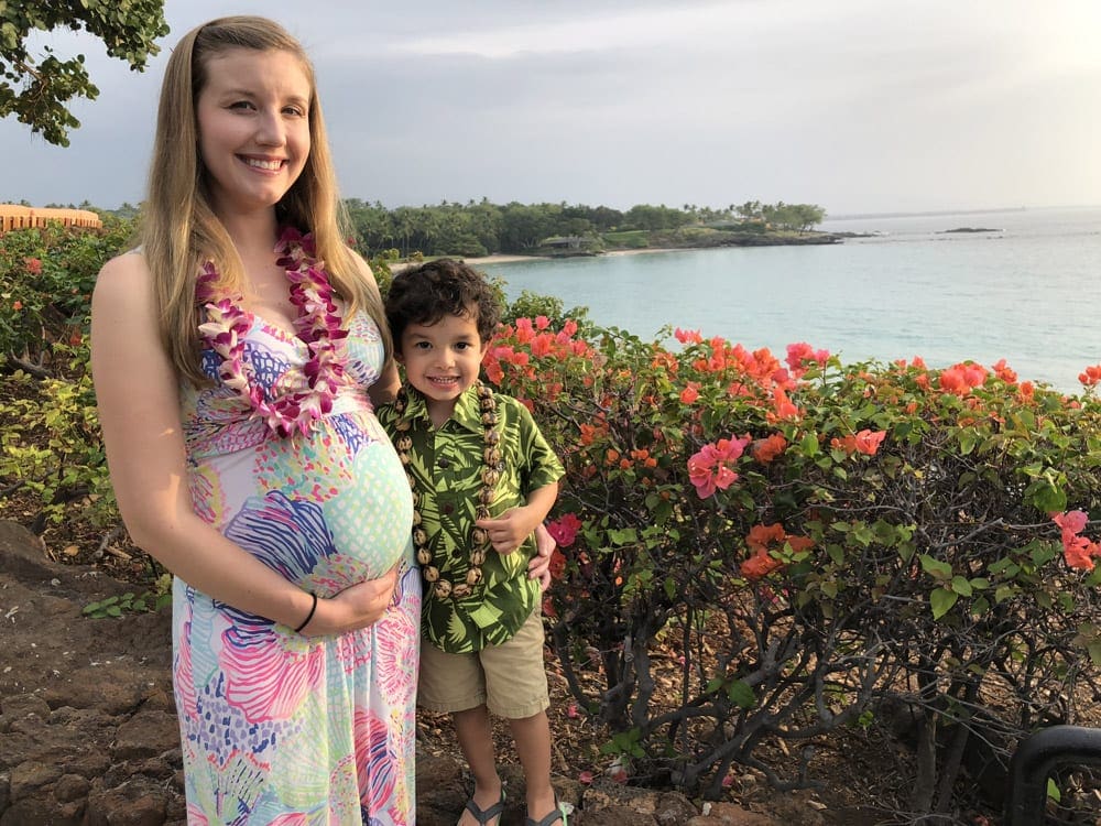 A pregnant mom holds her tummy, while standing next to her toddler son on a beautiful day along the water in Maui, one of the best spring break destinations for families around the world.