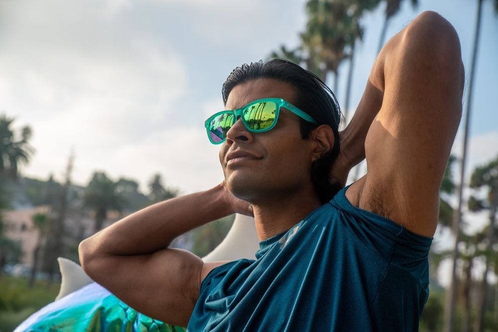 A man stretches with his hands behind his head while wearing goodr polarized sunglasses on a sunny day.