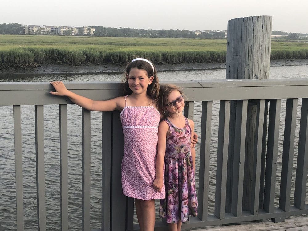 Two girls, both wearing pink dresses, smile while posing on a boardwalk while vacationing in Hilton Head.