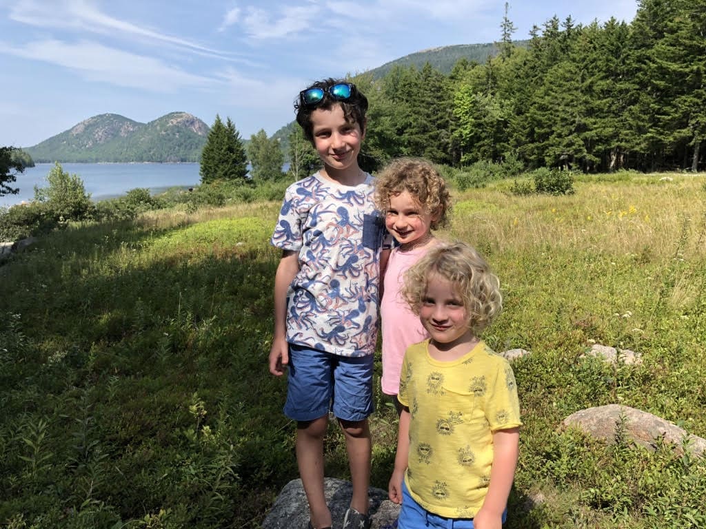 Three kids stand side by side, smiling, on a sunny day while vacationing in Maine.