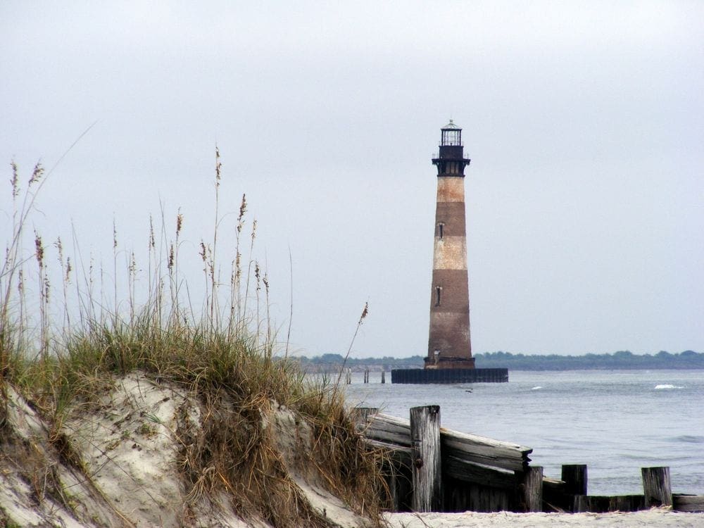 Across a grassy beach hill, the Morris Island light house is seen on an overcast day.