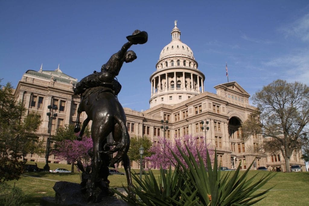 An image of the Texas Capitol Building and its manicured gardens, eating a black statue in the foreground. 