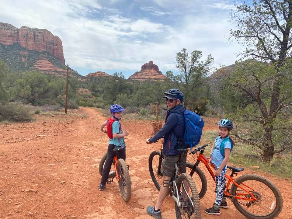 A dad and two kids perched on bikes look behind them at the camera while the iconic Sedona red rocks rest in the distance.