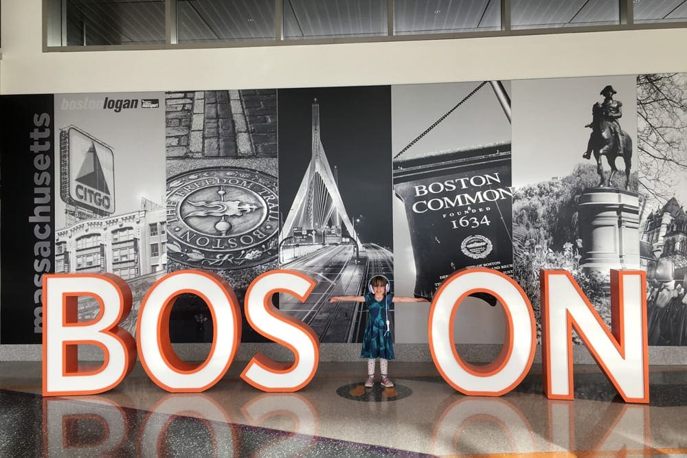 A young child uses their arms to make the "T" in a large sign reading "BOSTON".