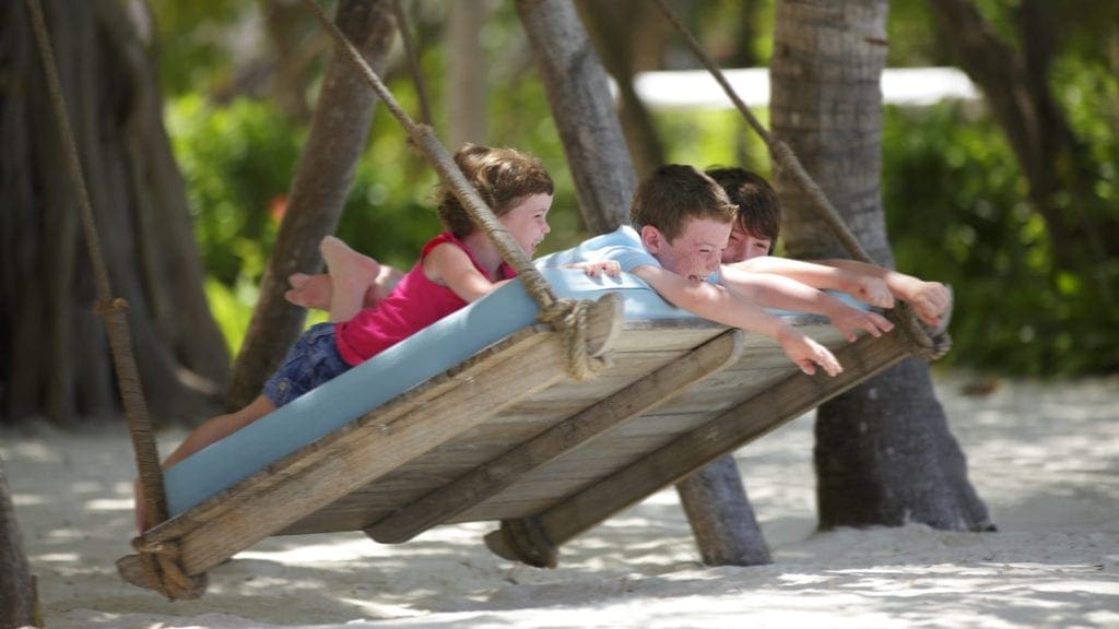 Three kids swing on a huge beach-side swing at the Four Seasons Resort Maldives At Kuda Huraa.