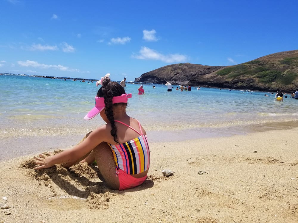 A young girl wearing a brightly striped swim suit and pink vizor sits on Hanauma Bay Beach in Oahu, one of the Hanauma Bay Nature Preserve with kids.