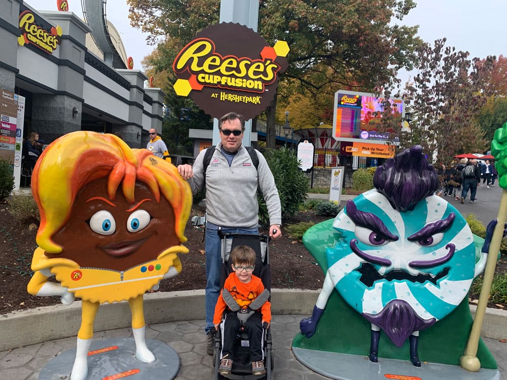 A dad stands behind his son, sitting in a stroller, between two mascots at Hershey Park.