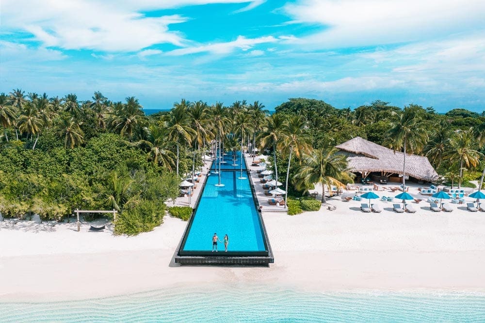 A couple stands at the edge of a large pool surrounded by lush palms at the Hotel Fairmont Maldives - Sirru Fen Fushi, one of the best family hotels in the Maldives.