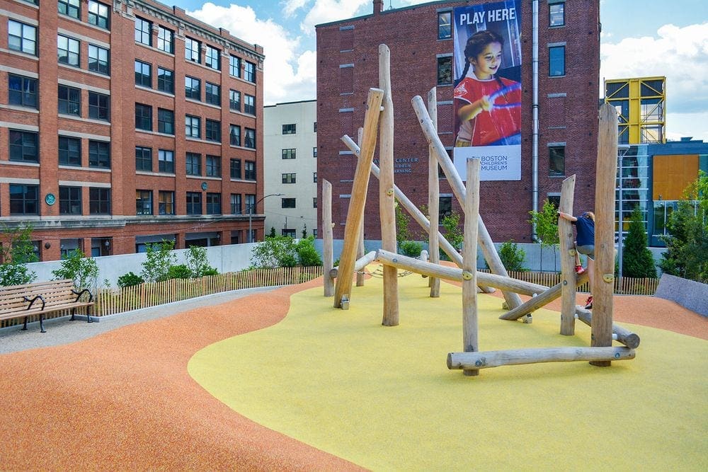 A large wooden play structure in Martin's Park, near the Boston Children's Museum, surrounded by large buildings.