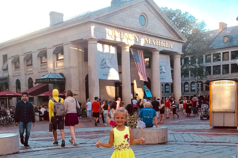 A young girl wearing a yellow dress and a huge smile stands outside of the Quincy Market.