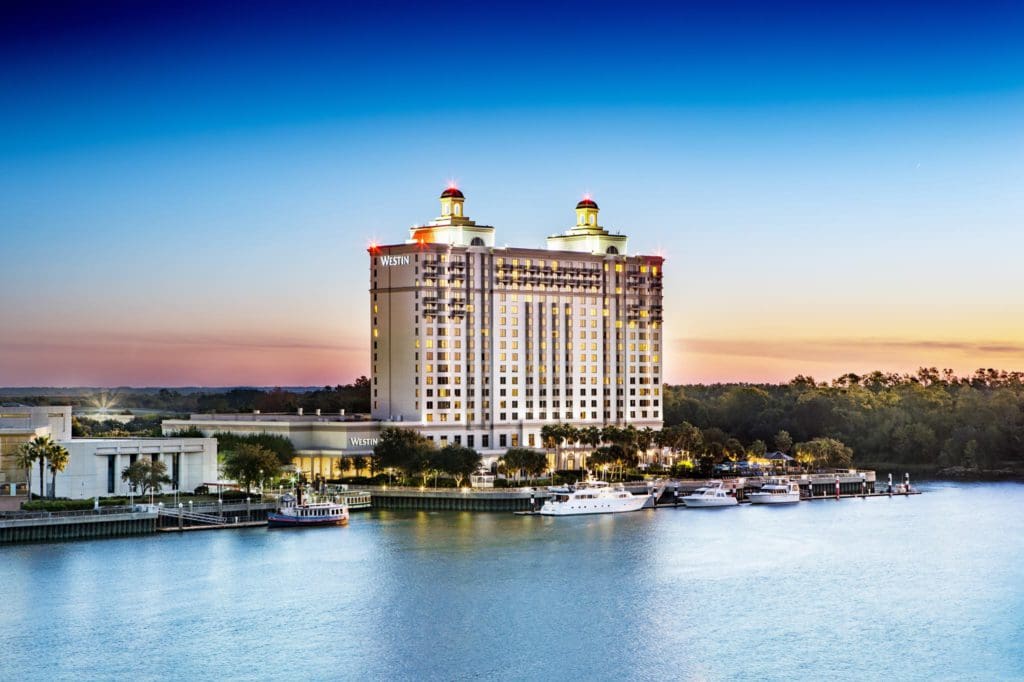 A view of the The Westin Savannah Harbor Golf Resort & Spa at sunset, beyond the water and marina, one of the best hotels in Savannah for families.