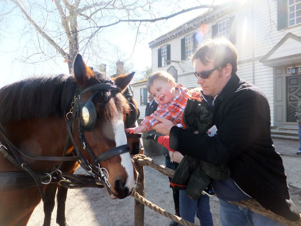 A dad holds his infant son, who leans over to pet a horse while they explore Williamsburg, Virgina, one of the best weekend getaways near DC for families.