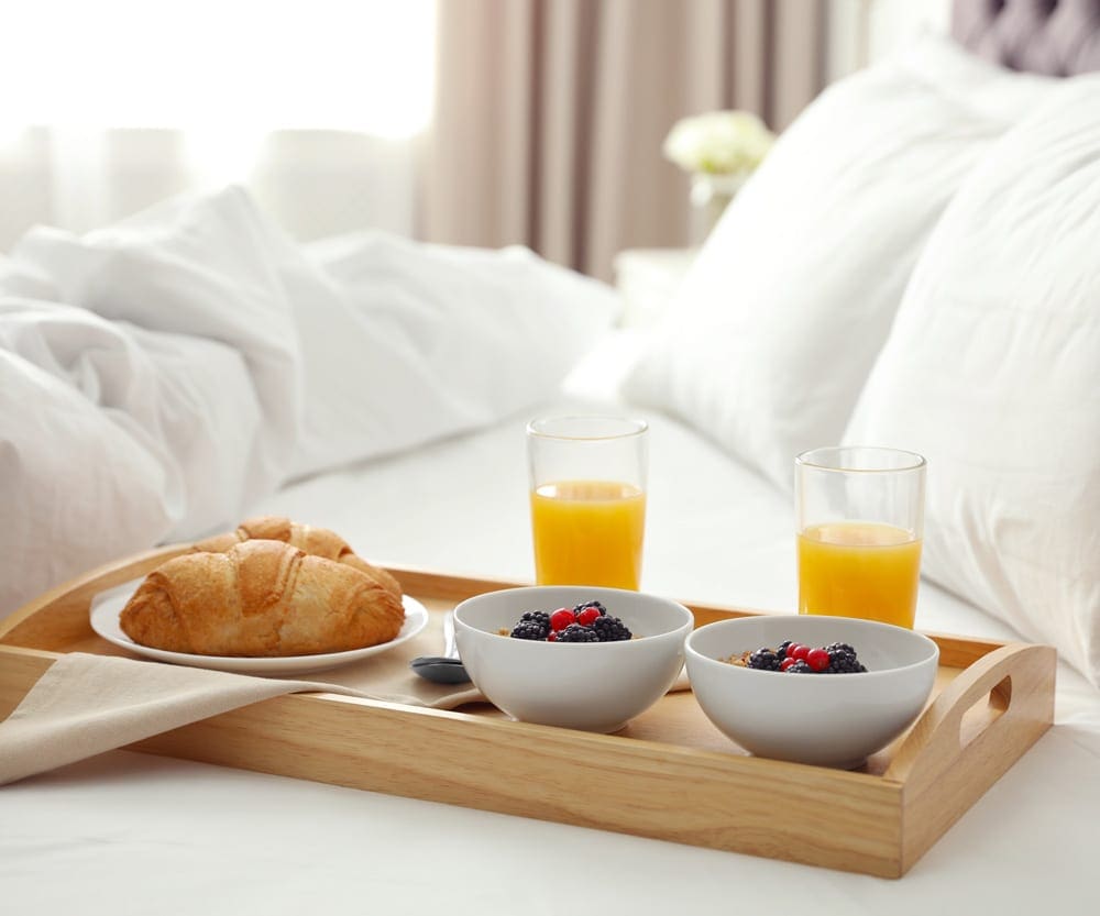 A breakfast in bed tray holds two orange juices, two cups of fruit, and a croissant on a lush, crisp white bed at a hotel.