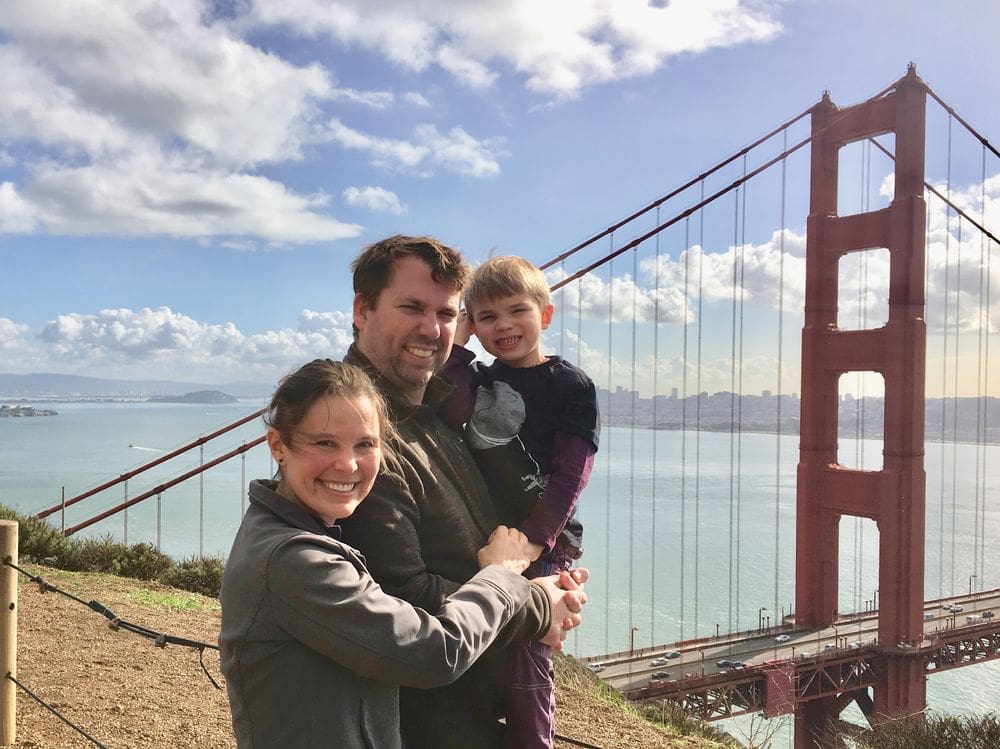 A family of three stands together with the Golden Gate Bridge and the San Francisco Bay behind them.