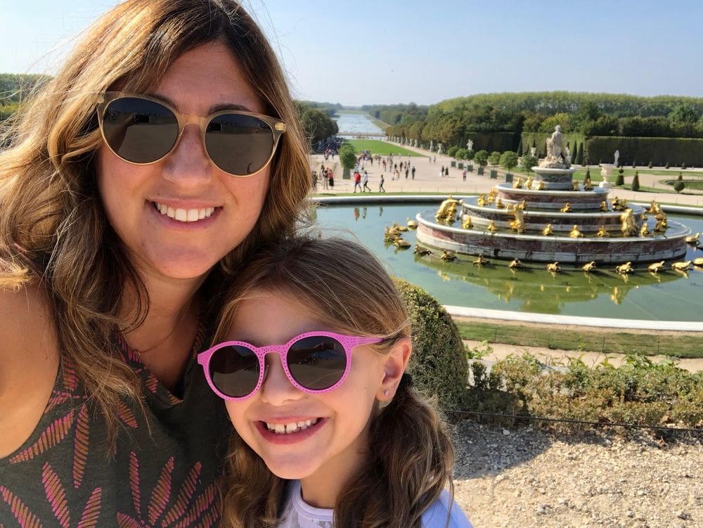 A mom and her daughter snap a selfie with the fountains of the Palace of Versailles behind them.