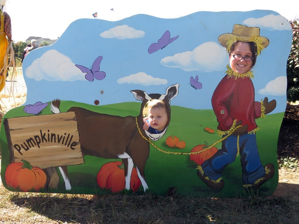 A mom and son poke their heads out of a cardboard cutout sign featuring a farmer and donkey at Pumpkin Village.