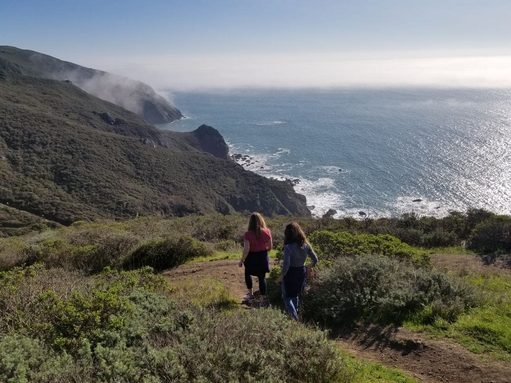 Two girls hike down a trail with an overlook of the ocean near Marin Headlands.
