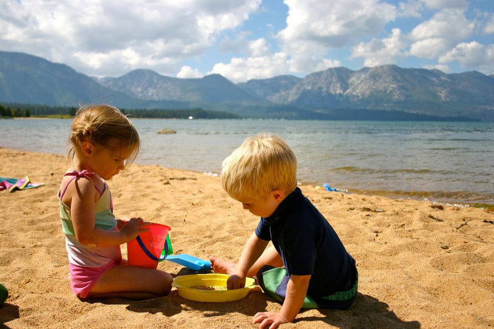 Two toddlers play in the sand near Lake Tahoe with mountains in the distance.
