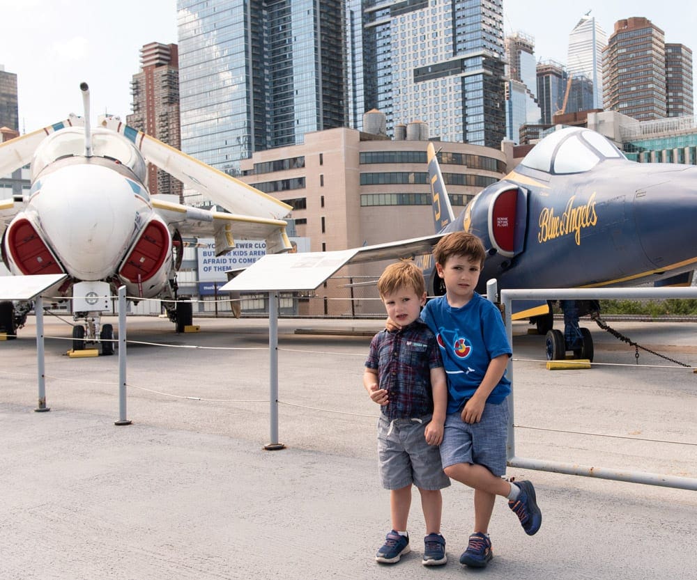 Two young boys standing in front of the Intrepid Air & Space Museum with airplanes in the background and a view of the Manhattan skyline.