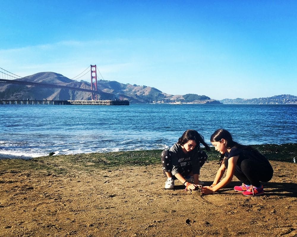 Two girls play in the sand with San Francisco Bay in the background.